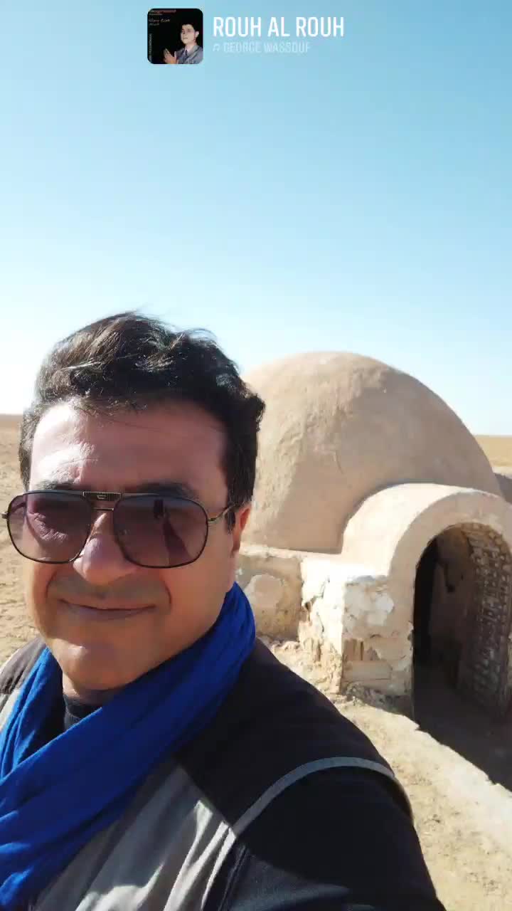 Salah, guide BHS Travel, dans le désert avec lunettes de soleil et foulard bleu, une bâtisse en dôme traditionnelle en arrière-plan.